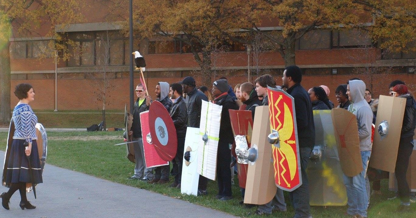 On the left, a short lady in a snazzy blue and white sweater faces a group of students holding cardboard Roman-style shields and a stuffed eagle on a stick.