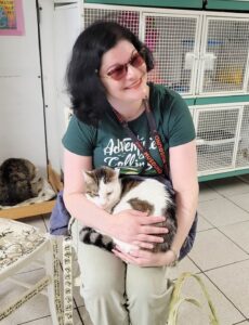 A medium-sized pale lady with black bobbed hair and sunglasses sits grinning on a chair in a room full of kennels. There is a majestic Roman cat named Zeyu on her lap, and he purrs very loudly. Both the cat and professor lady are smol.
